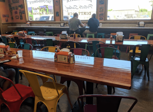 A casual dining area with colorful chairs and a long wooden table, featuring two people sitting by the window.