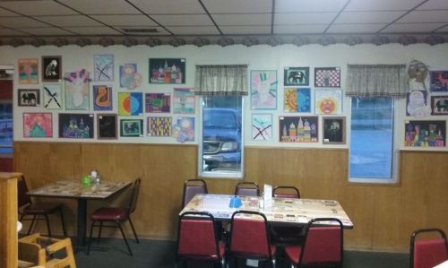 A cozy restaurant interior featuring a wall adorned with colorful children's artwork and empty tables with chairs.