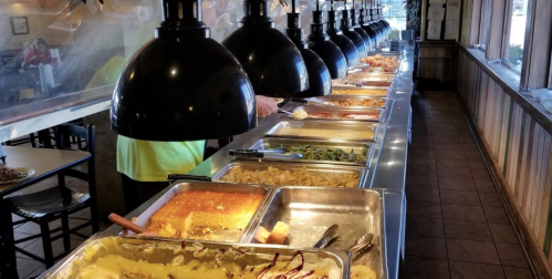 A buffet line with various dishes under warmers, featuring trays of food and overhead lighting in a casual dining setting.
