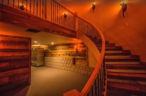 A warmly lit staircase with wooden railings curves around a cozy, rustic hallway featuring stone walls.