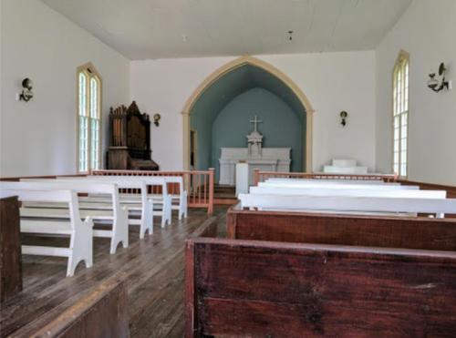 Interior of a simple church with wooden pews, an altar, and a large arched window in a bright, airy space.