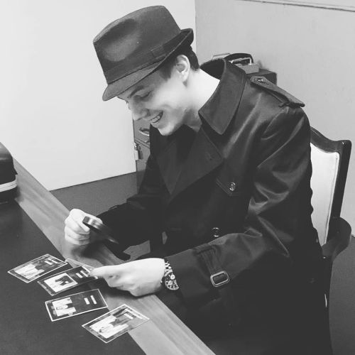 A person in a fedora and trench coat examines photos on a table, smiling with a focused expression.