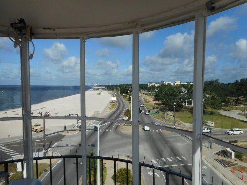 View from a lighthouse showing a beach, road, and trees under a partly cloudy sky.