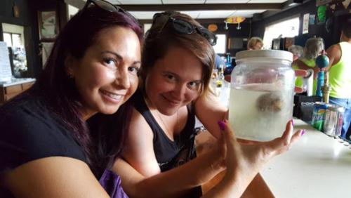Two women smile while holding a jar of liquid at a bar, with people in the background.