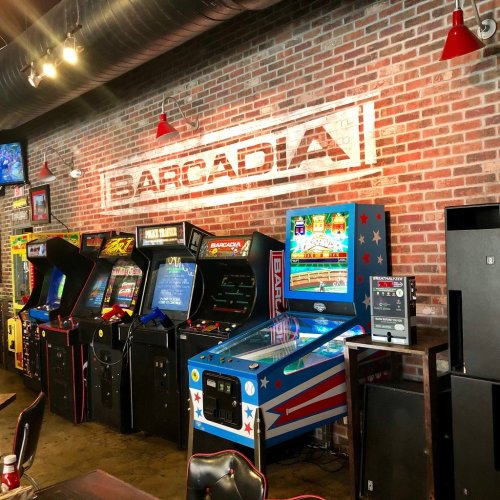 A wall of vintage arcade games and a pinball machine in a bar setting, with a brick backdrop and "Barcadia" sign.