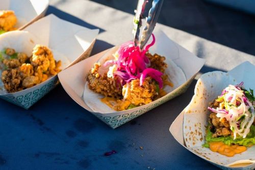 Three tacos on a table, one being topped with pink pickled onions and slaw, featuring crispy fried filling.