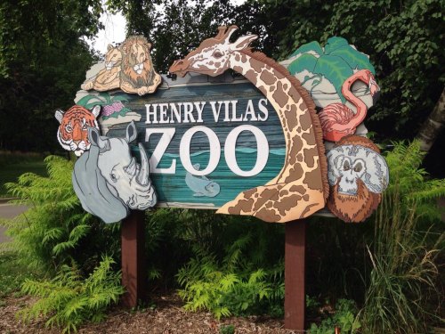 Colorful sign for Henry Vilas Zoo featuring animal illustrations like a lion, giraffe, and flamingo, surrounded by greenery.