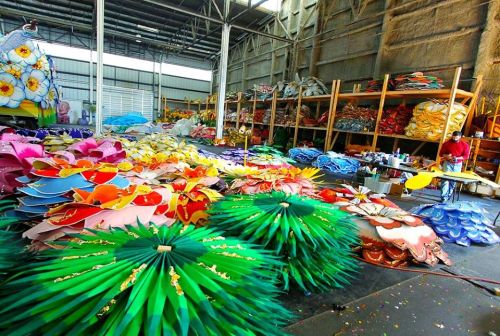 A vibrant workshop filled with colorful paper flowers and decorations, with a person working at a table in the background.