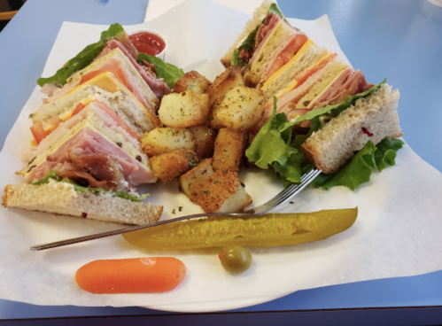 A plate with a club sandwich, crispy potatoes, a pickle, and a carrot and olive garnish on a blue table.