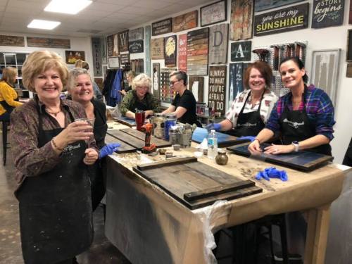 A group of women smiling and working on crafts at a workshop, with tools and materials on a table.