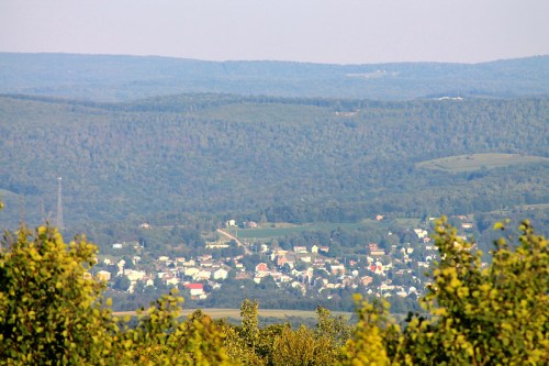 A panoramic view of a small town nestled in green hills, surrounded by trees and distant mountains under a clear sky.