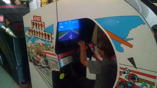 A child plays an Atari racing arcade game, gripping the steering wheel and focused on the screen displaying a racetrack.