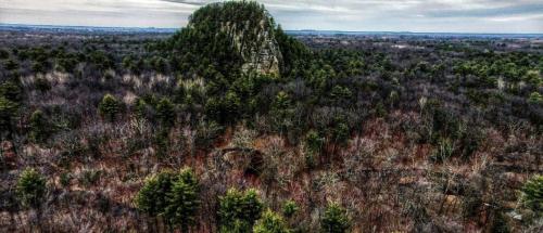 Aerial view of a rocky outcrop surrounded by dense, leafless trees and a cloudy sky in the background.