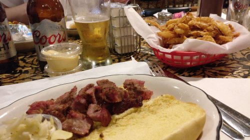 A plate with sliced meat, a piece of bread, and a side of coleslaw, with beer and onion rings in the background.