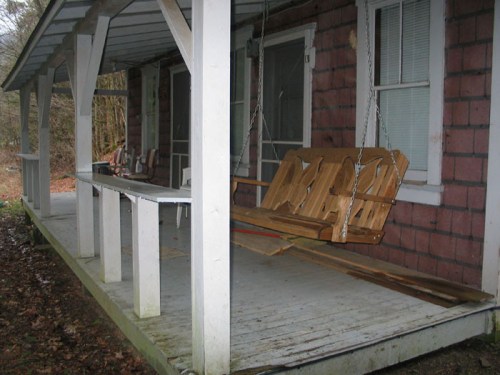 A wooden porch with a swing, surrounded by trees, featuring a weathered floor and a few chairs in the background.