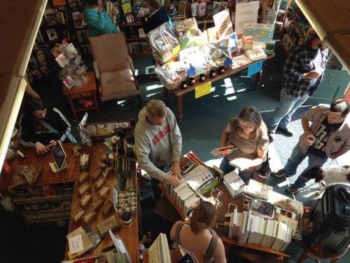 Aerial view of a busy bookstore with people browsing books and interacting at tables. Sunlight streams in through windows.