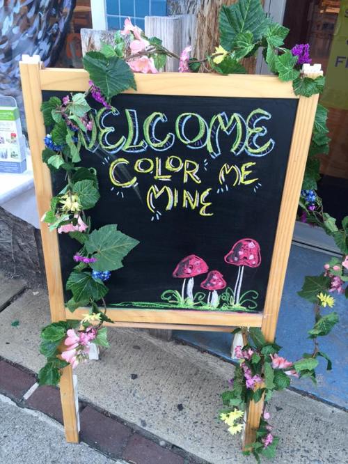 A chalkboard sign reading "Welcome Color Me Mine," decorated with flowers and mushrooms.