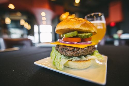 A close-up of a juicy burger with cheese, lettuce, tomato, pickles, and onion on a plate, with a glass of beer in the background.