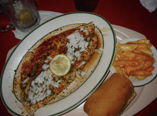 A plate featuring a baked fish topped with herbs and lemon, served with macaroni and cheese and a bread roll.