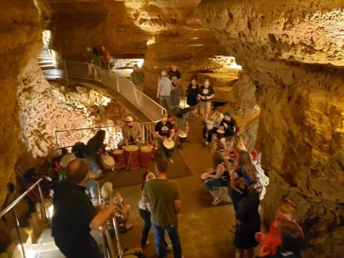 A group of people gathered in a cave, some playing drums, others sitting on the floor, with rock formations around them.