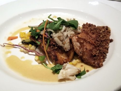 A plated dish featuring grilled fish, crab, and a corn salad, garnished with greens and a light sauce.