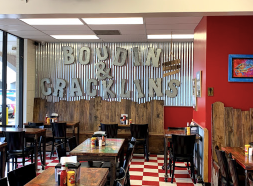Interior of a restaurant featuring a "Boudin & Cracklins" sign, wooden tables, and a red accent wall.
