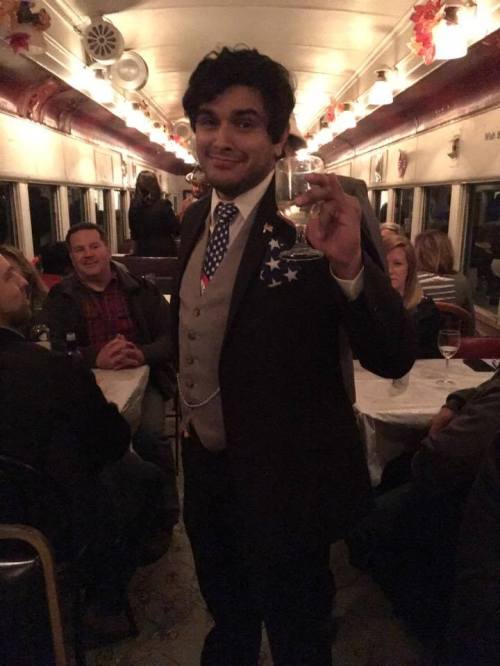 A man in a suit with an American flag pin holds a drink, smiling in a festive train car filled with people.