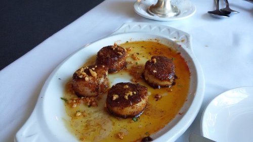 A white dish with four seared scallops topped with garlic, sitting in a flavorful sauce.