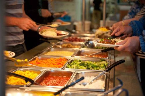 A buffet with various food options, including vegetables, pasta, and sauces, as people serve themselves.