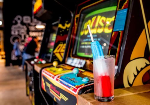 A colorful drink with a blue and red popsicle sits in front of vintage arcade game machines.