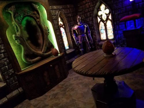 A dimly lit medieval room featuring a suit of armor, a round wooden table, and a large, dark mirror with green lighting.