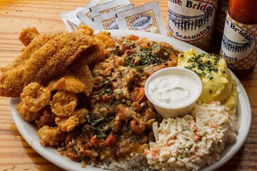 A plate of fried fish, shrimp, crawfish étouffée, mashed potatoes, coleslaw, and dipping sauce, with seasoning packets in the background.