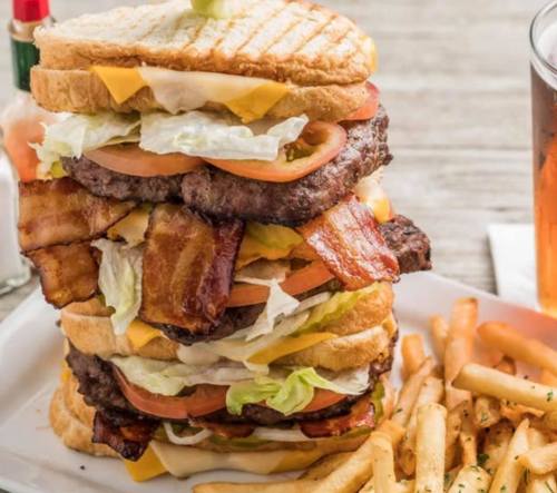 A towering sandwich with layers of beef, bacon, cheese, lettuce, and tomato, served with fries and a drink.