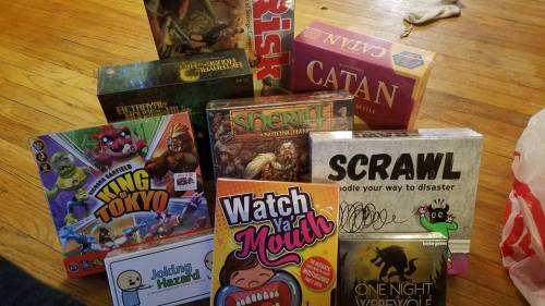 A collection of board games stacked on a wooden floor, including Catan, King of Tokyo, and Betrayal at House on the Hill.