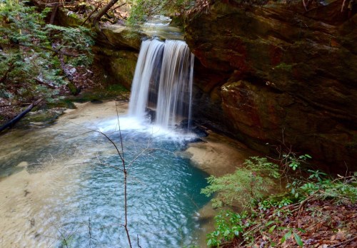 A serene waterfall cascades into a clear pool surrounded by lush greenery and rocky terrain.