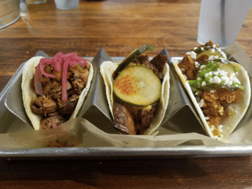 Three tacos on a tray: one with pulled meat and pickled onions, one with grilled vegetables, and one with spicy chicken.