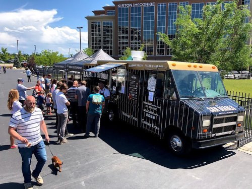 A food truck with a long line of people waiting outside, set against a backdrop of a modern building and blue sky.