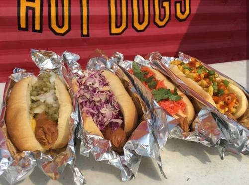 Four gourmet hot dogs in buns, each topped with different colorful ingredients, wrapped in foil.