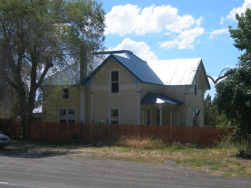 A yellow Victorian-style house with a metal roof, surrounded by trees and a wooden fence under a blue sky.
