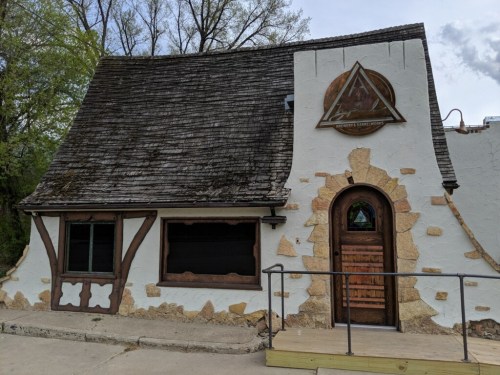 A unique, rustic building with a sloped roof, stone accents, and a triangular emblem on the facade.
