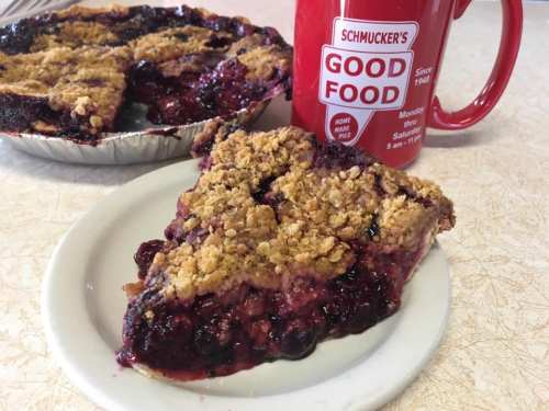 A slice of berry pie with a crumb topping on a plate, next to a red mug that says "Good Food."
