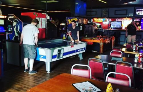 Two people play air hockey in a retro arcade filled with games and tables.
