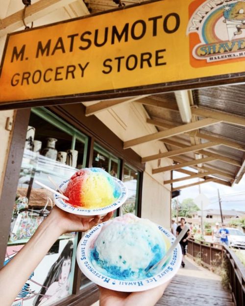 Two hands hold colorful shaved ice bowls in front of M. Matsumoto Grocery Store sign in Hawaii.