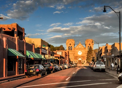 A street view of a charming town with adobe buildings, shops, and a church in the background under a cloudy sky.