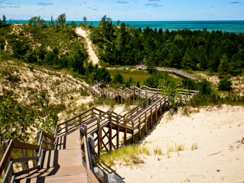 A winding wooden boardwalk through sandy dunes and greenery, leading to a view of the lake in the distance.