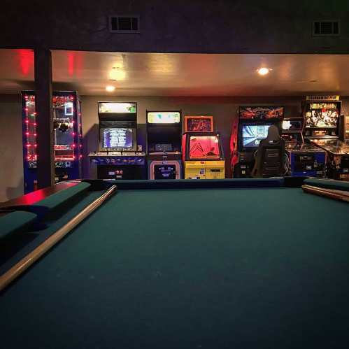 A pool table in the foreground with colorful arcade machines lined up in the background, illuminated by soft lights.