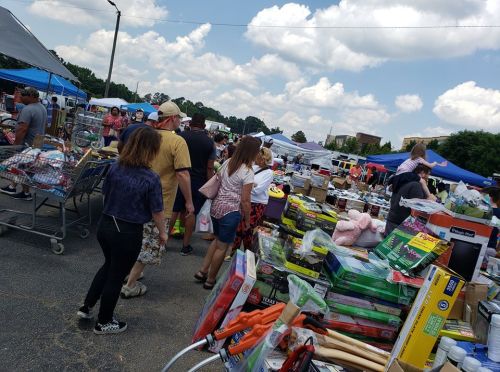 A bustling flea market scene with shoppers browsing various stalls and tables filled with items under blue tents.