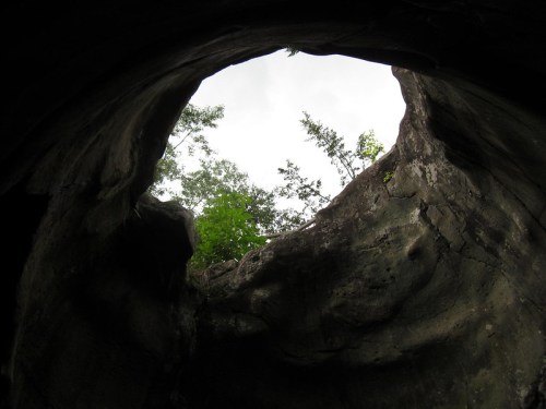 View from inside a cave looking up at a circular opening with trees and cloudy sky visible above.