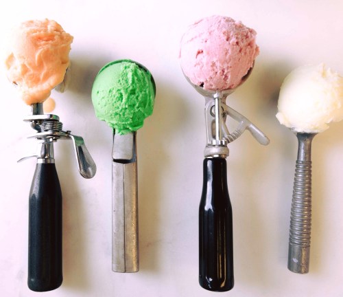 Four ice cream scoops in different colors—orange, green, pink, and white—on metal scoops against a white background.