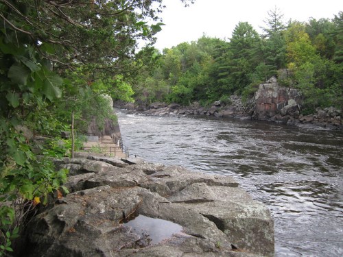 A serene river flows past rocky banks, surrounded by lush green trees and a peaceful natural landscape.